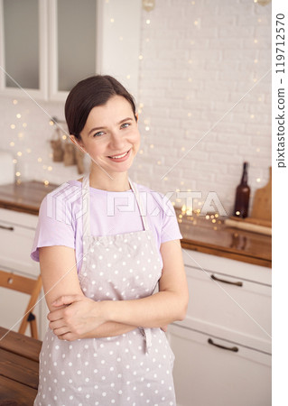 Smiling woman in polka dot apron standing in modern kitchen with warm ambient Smiling woman in polka dot apron standing in modern kitchen with warm ambient 119712570