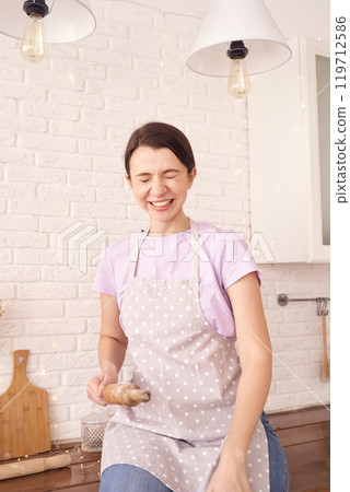 Joyful woman in apron holding ginger root in modern kitchen with white brick 119712586
