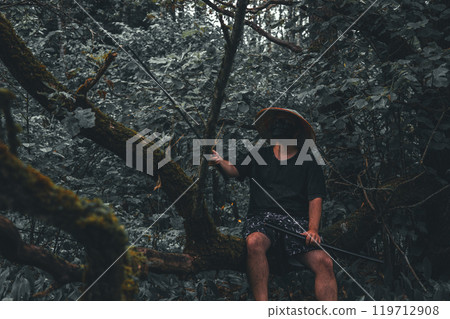 Warrior with katana in the forest. A mysterious man in a black mask and straw hat sits on a fallen tree in the forest, holding a katana, blending into the shadows, creating intrigue. 119712908