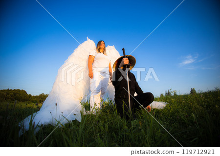 Angel spreads wings protecting samurai with katana An angel with large white wings and a samurai holding a katana pose together in a field, creating a striking juxtaposition against a clear blue sky. 119712921