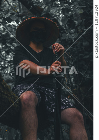 Cosplayer. Vertical. Man with katana sits majestically on tree trunk with katana. Samurai rests with katana in forest alone. 119712924