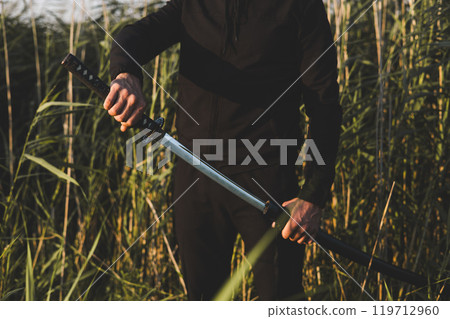 Close up of a martial artist unsheathing his katana sword in nature, surrounded by tall grass, with a focused and determined expression on his face. The man is training with a katana. Close up of a martial artist unsheathing his katana sword in nature, surrounded by tall grass, with a focused and determined expression on his face. The man is training with a katana. 119712960