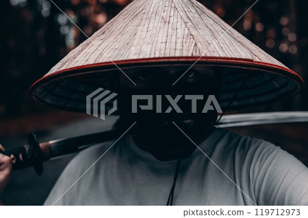 A mysterious man in a conical hat and mask confidently holds a katana in a serene forest, radiating strength and danger. A fearless samurai warrior stands with a katana on his shoulder. A mysterious man in a conical hat and mask confidently holds a katana in a serene forest, radiating strength and danger. A fearless samurai warrior stands with a katana on his shoulder. 119712973