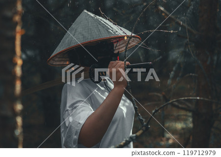 A warrior cosplayer stands in a rainy forest holding a katana sword. The mysterious figure exudes strength and power, evoking danger and excitement with a katana on his shoulder. A warrior cosplayer stands in a rainy forest holding a katana sword. The mysterious figure exudes strength and power, evoking danger and excitement with a katana on his shoulder. 119712979