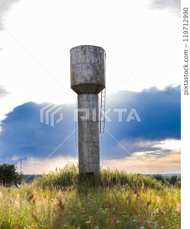 An old, time-worn water tower stands amidst grass and wildflowers against a cloudy sky at sunset. Beautiful water tower against a cloudy sky at sunset. An old, time-worn water tower stands amidst grass and wildflowers against a cloudy sky at sunset. Beautiful water tower against a cloudy sky at sunset. 119712990