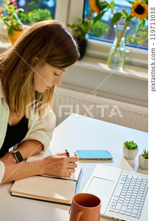 Woman working at home, writing in notebook and using laptop Woman working at home, writing in notebook and using laptop 119713038