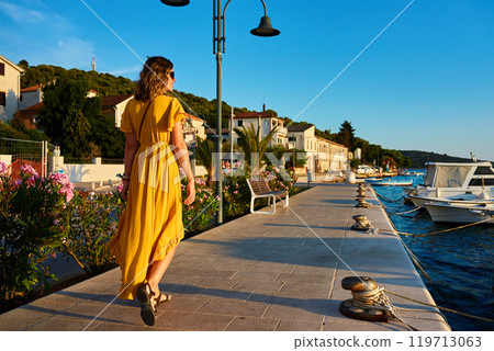 Woman in yellow dress walking down sunny promenade in coastal town Woman in yellow dress walking down sunny promenade in coastal town 119713063