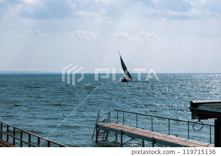 Seascape with sailing boat in the distance and pier Seascape with sailing boat in the distance and pier 119715136