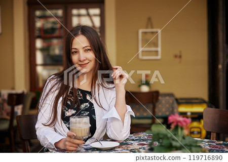 coffee cup, Relaxed Woman, Morning Coffee. At charming outdoor cafe, young woman smiles while holding coffee cup, creating welcoming and relaxed atmosphere. 119716890