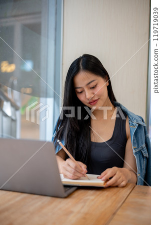 A woman taking notes in a book while studying online or working remotely on her laptop at a cafe. 119719039
