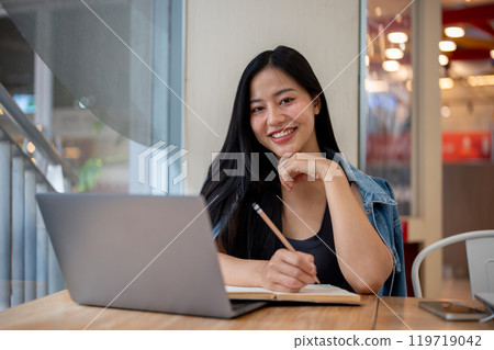 A woman smiling at the camera while studying online or working remotely on her laptop at a cafe. A woman smiling at the camera while studying online or working remotely on her laptop at a cafe. 119719042
