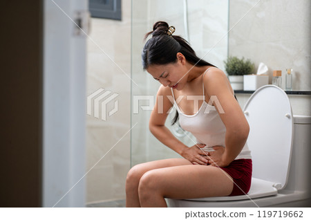 An Asian woman in pajamas sitting on a toilet, gently touching her stomach, severe stomach pain. An Asian woman in pajamas sitting on a toilet, gently touching her stomach, severe stomach pain. 119719662