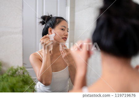 A beautiful Asian woman is massaging her face with a Gua Sha stone scraper in front of the mirror. A beautiful Asian woman is massaging her face with a Gua Sha stone scraper in front of the mirror. 119719676