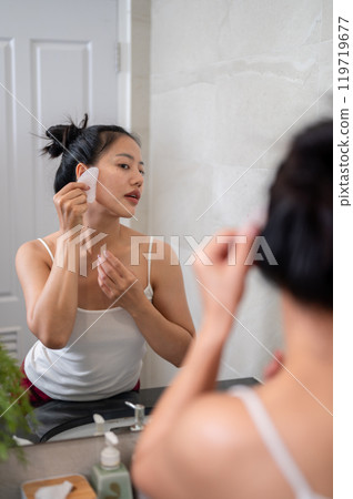 A beautiful Asian woman is massaging her face with a Gua Sha stone scraper in front of the mirror. 119719677