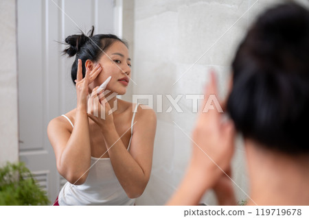 A beautiful Asian woman is massaging her face with a Gua Sha stone scraper in front of the mirror. A beautiful Asian woman is massaging her face with a Gua Sha stone scraper in front of the mirror. 119719678