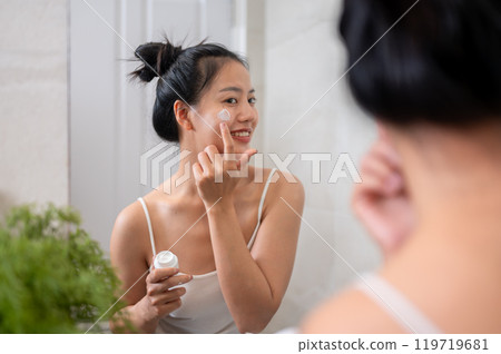 A natural Asian woman is applying facial cream in front of the mirror in the bathroom after shower. A natural Asian woman is applying facial cream in front of the mirror in the bathroom after shower. 119719681
