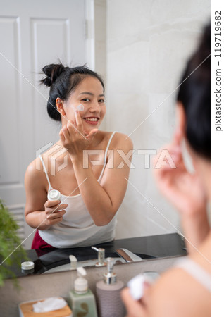A natural Asian woman is applying facial cream in front of the mirror in the bathroom after shower. 119719682
