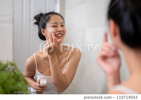 An attractive Asian woman is smiling at the mirror while applying facial cream to her face. 119719683