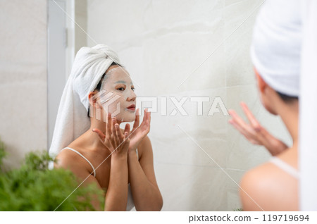 An Asian woman is applying a facial mask pad to her face in front of the bathroom mirror. 119719694