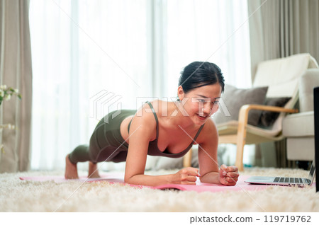A sporty, fit Asian woman is pranking on a yoga mat, practicing yoga in the living room. A sporty, fit Asian woman is pranking on a yoga mat, practicing yoga in the living room. 119719762