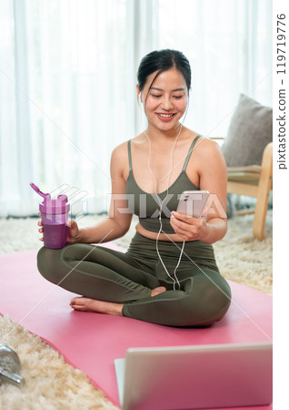 A beautiful, positive Asian woman in sportswear is resting on a yoga mat after her workout at home. A beautiful, positive Asian woman in sportswear is resting on a yoga mat after her workout at home. 119719776