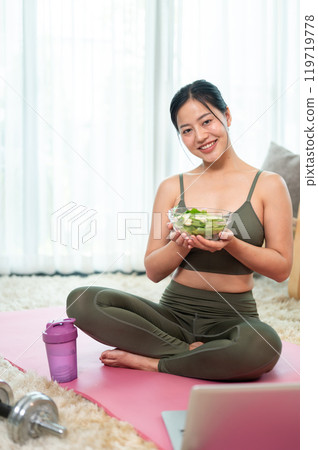 A healthy Asian woman is sitting on a yoga mat in the living room with a bowl of salad in her hand. A healthy Asian woman is sitting on a yoga mat in the living room with a bowl of salad in her hand. 119719778