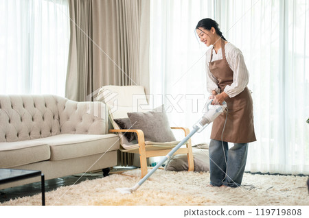 A beautiful Asian woman is cleaning a carpet with a vacuum cleaner in the living room at home. 119719808