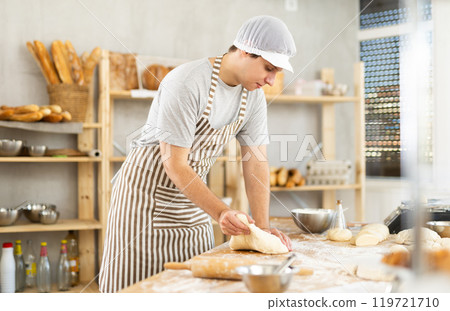 Portrait of confident professional hispanic young baker in apron kneading and working with raw yeasted dough in bakery 119721710