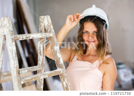 Young attractive girl in beige dress and protective helmet poses on stepladder in room being renovated Young attractive girl in beige dress and protective helmet poses on stepladder in room being renovated 119721730
