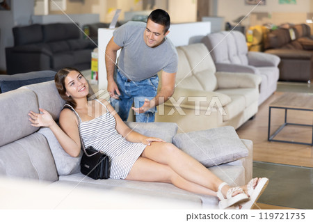 Couple chooses furniture in store.Wife is sitting on sofa,husband is standing next to her Couple chooses furniture in store.Wife is sitting on sofa,husband is standing next to her 119721735