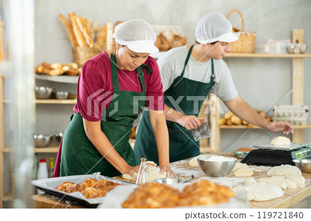 Skilled two baker in apron, adult female and young guy kneading dough, cutting and weighing portioned pieces on floured surface before baking Skilled two baker in apron, adult female and young guy kneading dough, cutting and weighing portioned pieces on floured surface before baking 119721840
