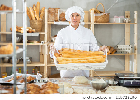 In working room of bakery, there is woman with baguettes in wicker basket in hands 119722190