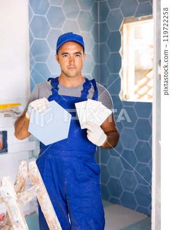 Skilled builder in work clothes posing with ceramic tiles and construction tools while repair and decoration in apartment 119722258