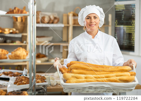 In working room of bakery, there is woman with baguettes in wicker basket in hands In working room of bakery, there is woman with baguettes in wicker basket in hands 119722284