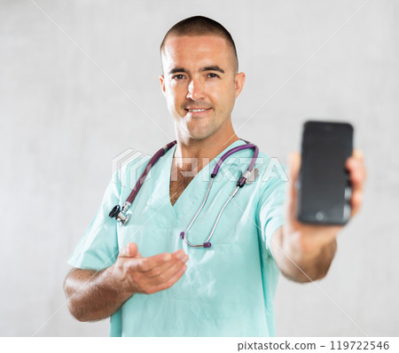 Male doctor in medical uniform posing with mobile phone in medical room Male doctor in medical uniform posing with mobile phone in medical room 119722546