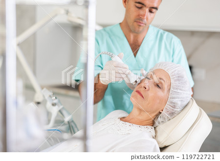 Cosmetologist performing anti-aging face hardware therapy procedure for aged female client in clinic of aesthetic medicine Cosmetologist performing anti-aging face hardware therapy procedure for aged female client in clinic of aesthetic medicine 119722721