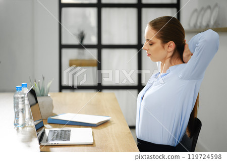 Woman stretching at a desk with a laptop during a work break in a modern office setting Woman stretching at a desk with a laptop during a work break in a modern office setting 119724598