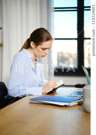 Young woman writing in a notebook at a desk by a window during daytime in a modern office Young woman writing in a notebook at a desk by a window during daytime in a modern office 119724601