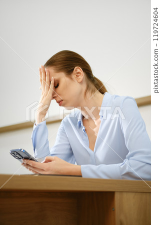 A woman sitting at a desk appears stressed while looking at her smartphone in a light-filled room 119724604