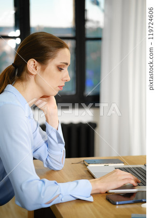 Professional woman typing on a laptop while sitting at a desk in a bright office environment 119724606
