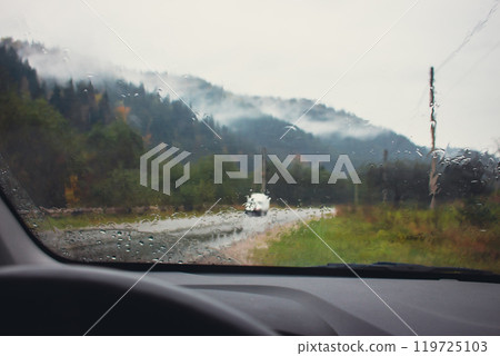 Driving in mountains on rainy day. Car dashboard with steering wheel with rain drops on glass and autumn mountains on background. Car on highway in mountains. Autumn vacations concept. Driving in mountains on rainy day. Car dashboard with steering wheel with rain drops on glass and autumn mountains on background. Car on highway in mountains. Autumn vacations concept. 119725103