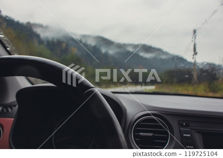 Driving in mountains on rainy day. Car dashboard with steering wheel with rain drops on glass and autumn mountains on background. Car on highway in mountains. Autumn vacations concept.  119725104