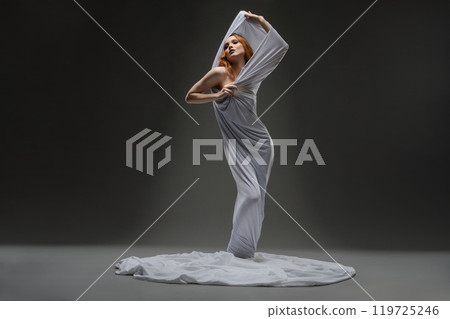 Elegant model in a flowing white dress poses dramatically in studio lighting 119725246
