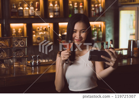 Asian woman taking a photo of herself while drinking whiskey at the bar. 119725307