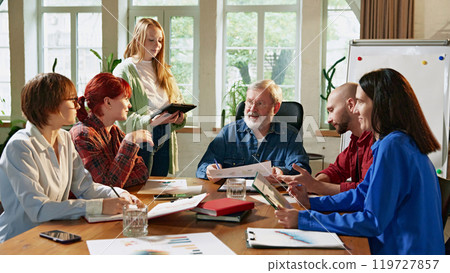 Team of diverse business professionals discussing strategies around conference table in modern, well-lit office. Modern workplace dynamics. 119727857