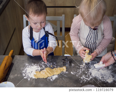 Two kids in aprons bake cookies in a kitchen, using cookie cutters to shape cookies from dough on a floured countertop. Laughter fills the room as they press, cut, and create cookies ready to bake. 119728079