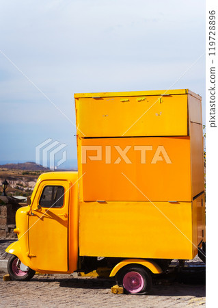 A yellow truck is parked by the road in front of a mountain 119728896