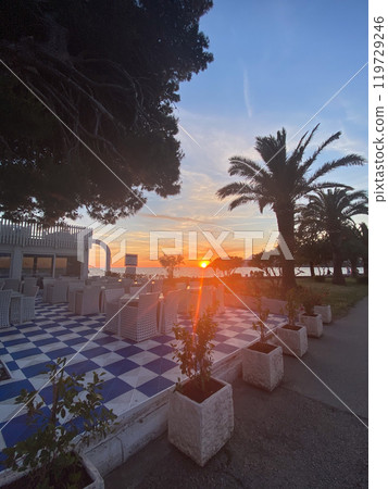 Vibrant sunset over a serene coastal terrace creates a picturesque dining atmosphere by the sea. The colorful sky enhances the peaceful setting, offering a perfect blend of nature and relaxation 119729246