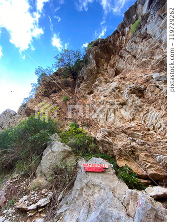 Rugged rock face rises sharply against a backdrop of blue sky dotted with white clouds, showcasing resilience of nature as a lone tree clings to cliffside. Contrast between the harshness of the rocks Rugged rock face rises sharply against a backdrop of blue sky dotted with white clouds, showcasing resilience of nature as a lone tree clings to cliffside. Contrast between the harshness of the rocks 119729262