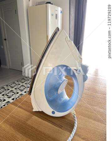 White and blue iron stands upright on a wooden table in a modern kitchen setting, capturing the essence of domestic chores. Natural light highlights the sleek design and functionality of the everyday 119729289